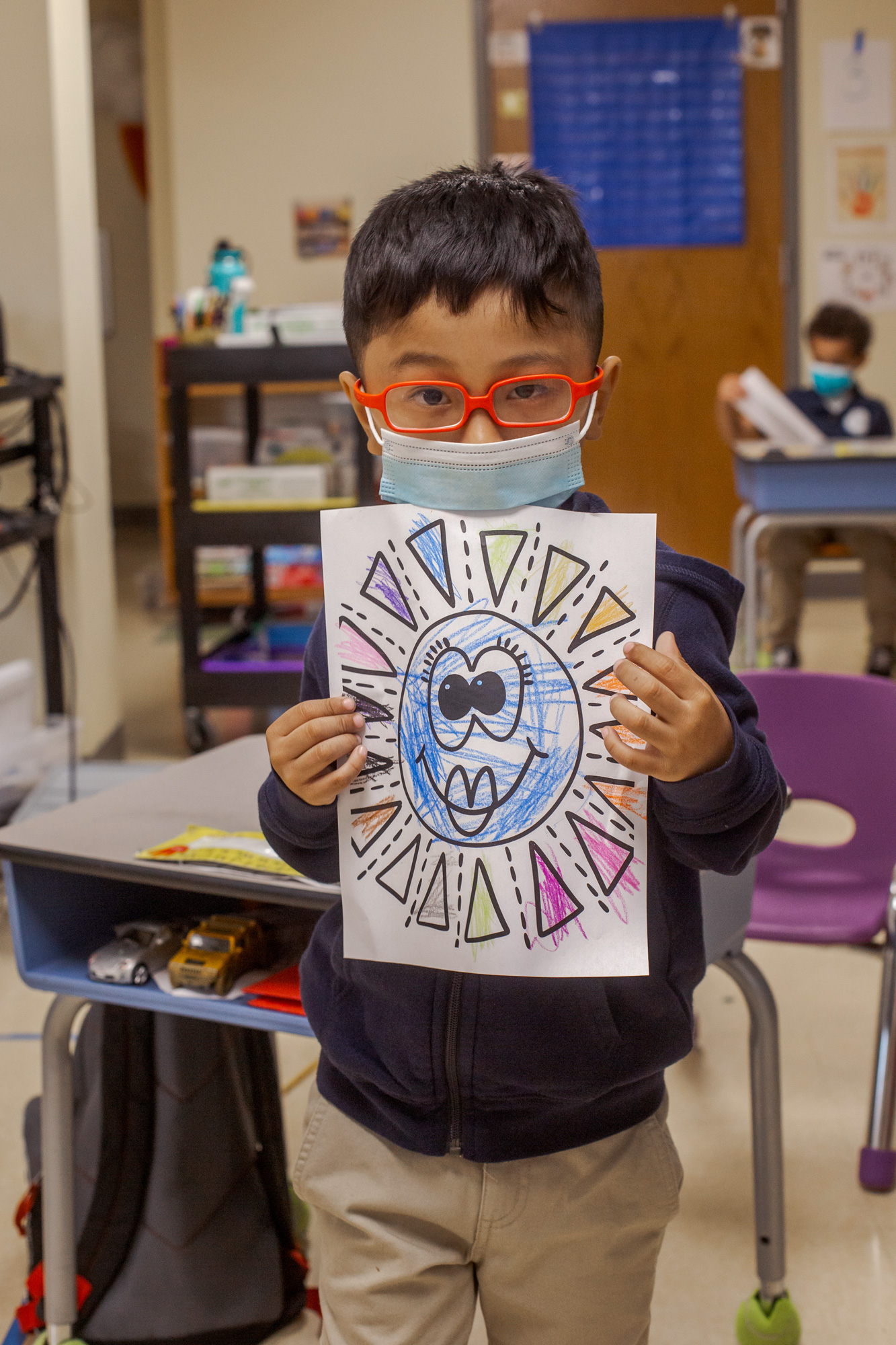 little boy with glasses and a mask on in a classroom holding up a coloring page
