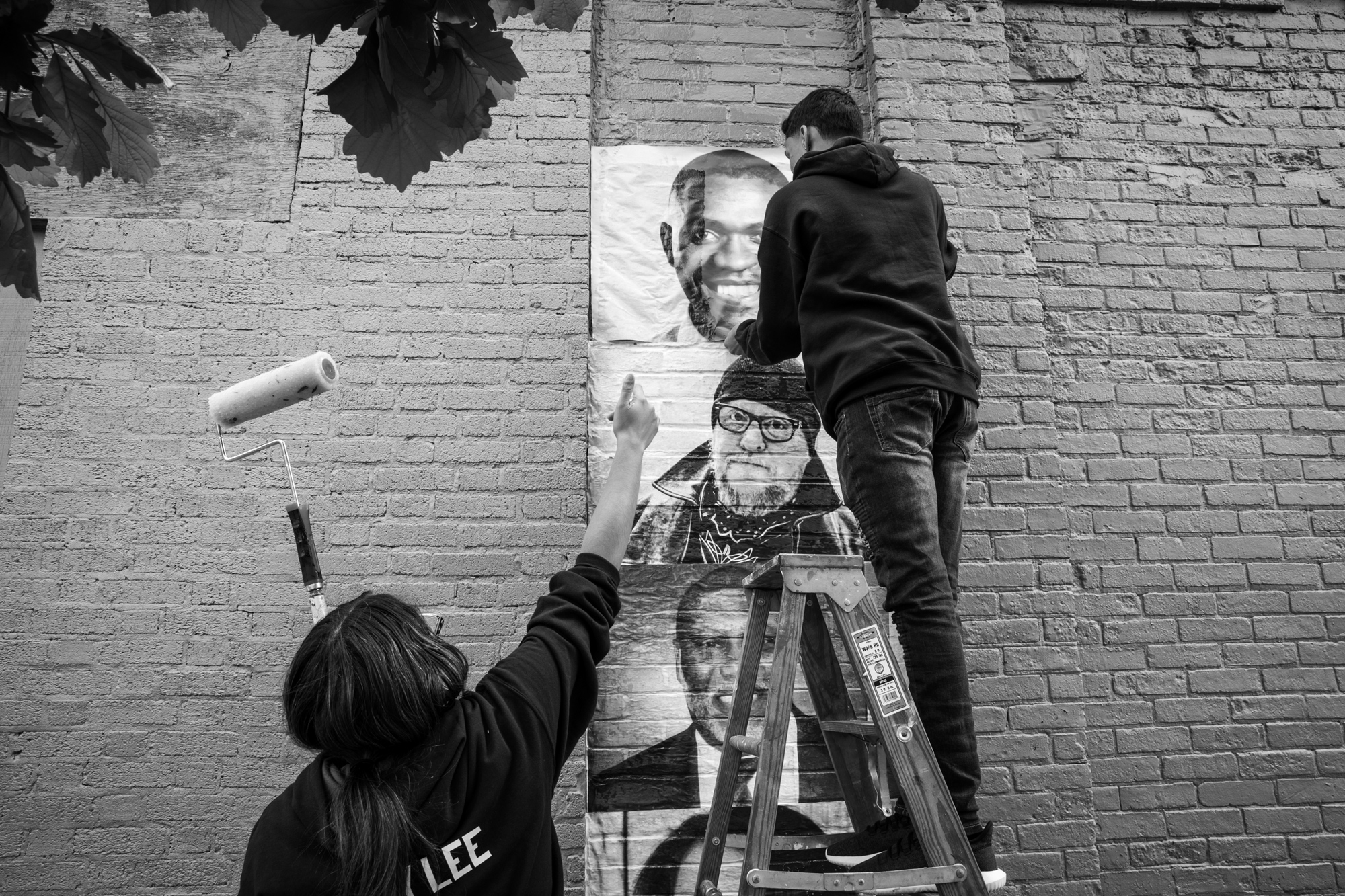 black and white photo of two people hanging people's portraits on the side of a brick building