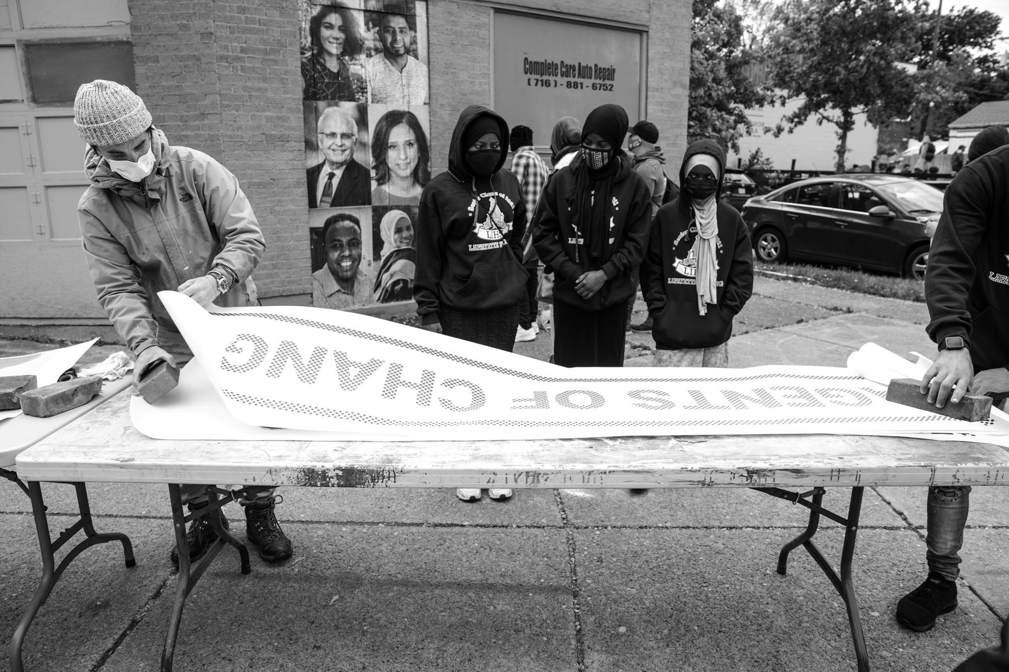 black and white photo of two people laying out the agents of change sign while people in the background watch