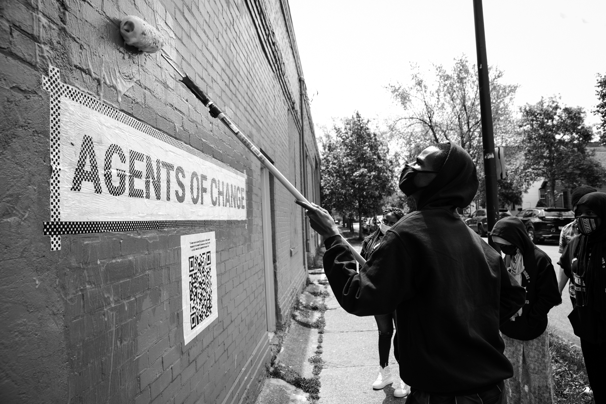 black and white photo of a group of people using a paint roller to hang the agents of change sign on the side of a brick building