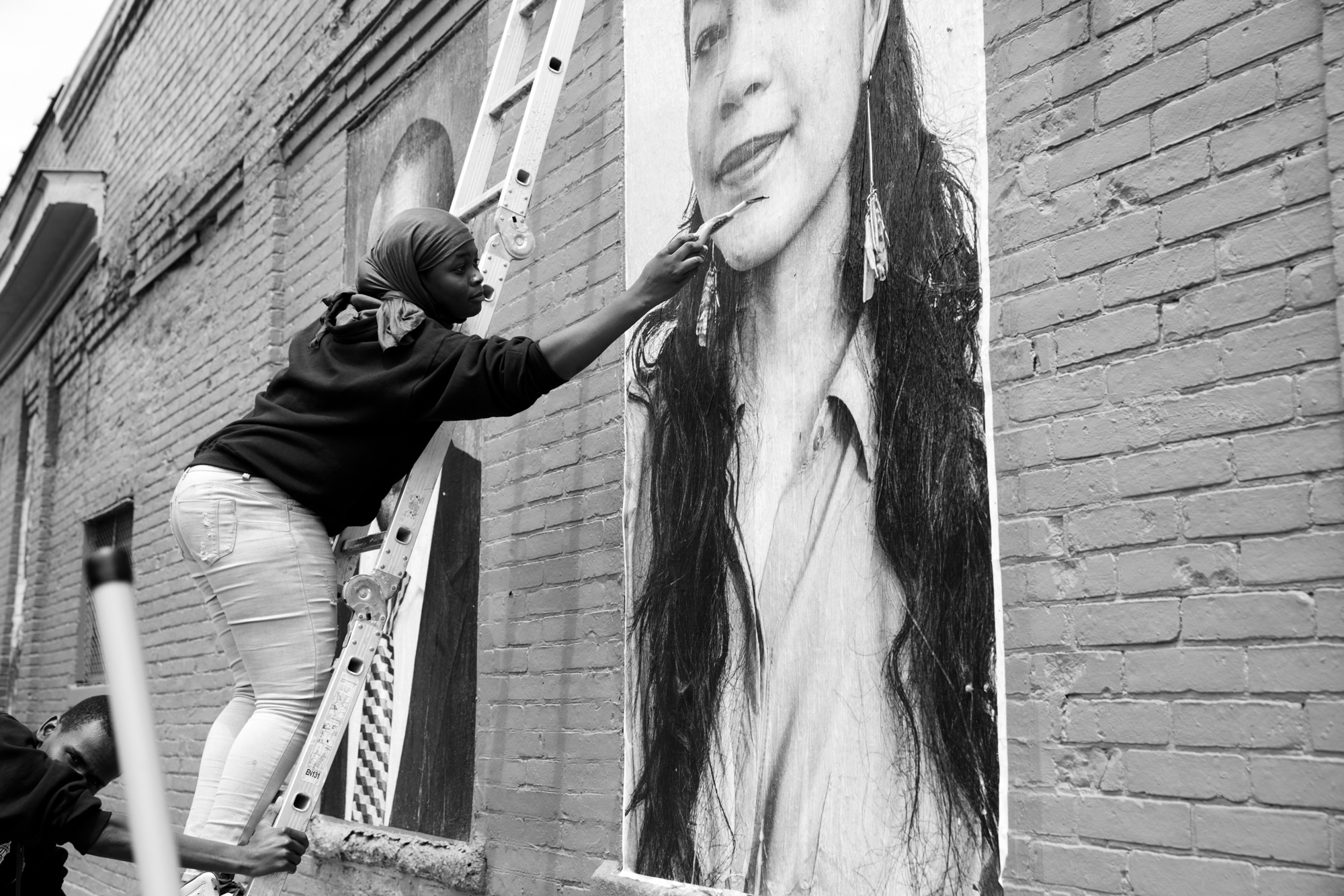 woman on a ladder that a man is stabilizing while she is putting the final touches on the person's portrait hanging on the side of a brick building