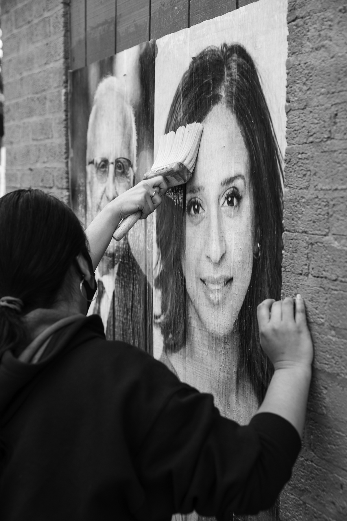 black and white photo of person using a paintbrush to hang woman's portrait on the side of a brick wall